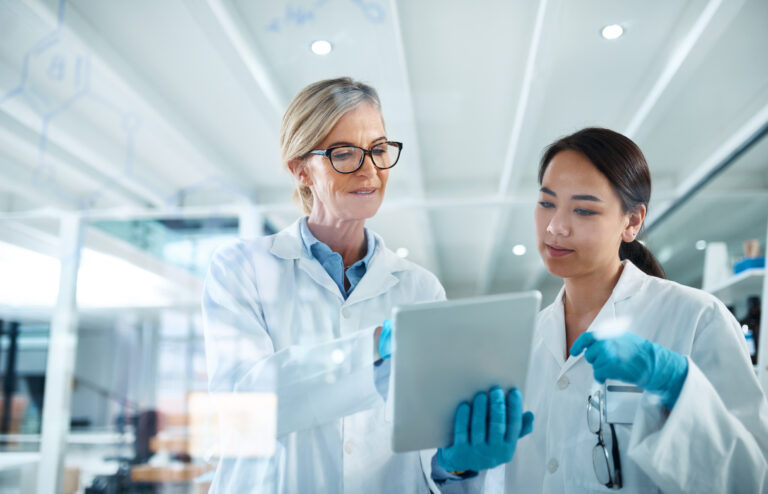 Deux scientifiques féminines, en blouse et équipées de gants, observent une tablette affichant des résultats de l'entreprise pharmaceutique où elles travaillent. La photo a été prise à travers une vitre de laboratoire.