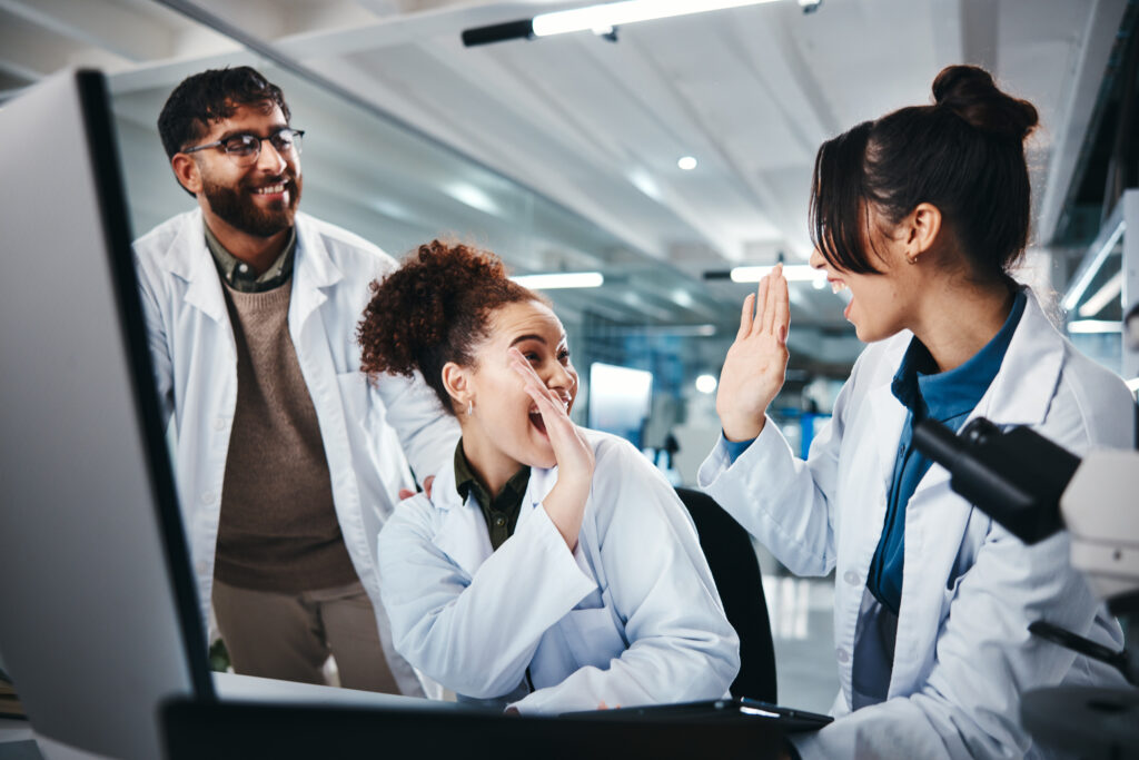 Trois scientifiques réunis devant un ordinateur dans un laboratoire. Un homme barbu portant des lunettes et une blouse blanche se tient debout, un sourire aux lèvres, observant avec bienveillance ses deux collègues. Les deux femmes, également en blouse blanche, échangent un high-five enthousiaste, exprimant leur joie face à une découverte ou un résultat prometteur.